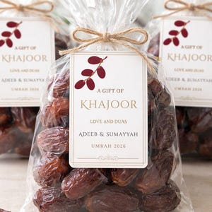 May include: Clear cellophane bags filled with dates, tied with twine and a tag. The tag reads "A Gift of Khajoor, Love and Duas, Adeeb & Sumayyah, Umrah 2026". The dates are a dark brown colour.