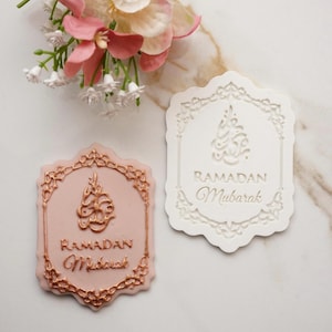 Peut inclure: Deux décorations Ramadan Moubarak. L'une est rose clair avec des accents dorés, l'autre est blanche. Les deux ont des bordures ornées et de la calligraphie arabe. Le texte "Ramadan Moubarak" est également présent. Des fleurs décoratives sont en arrière-plan.