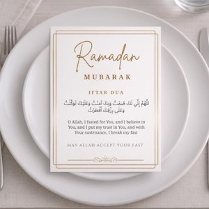 Puede incluir: Una tarjeta blanca con texto dorado y un borde dorado, colocada sobre un plato blanco. La tarjeta dice "Ramadan Mubarak" e incluye escritura árabe y texto en inglés para Iftar Dua, una oración para romper el ayuno. La tarjeta es parte de una mesa.