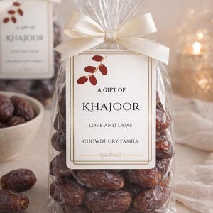 May include: A transparent cellophane bag filled with dates, tied with a cream ribbon. A rectangular tag with the text "A Gift of Khajoor, Love and Duas, Chowdhury Family" is attached. Dates are also in a white bowl.