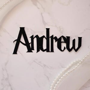Op de afbeelding: Een zwarte uitsparing van de naam "Andrew" in een gotisch lettertype, gecentreerd op een wit marmeren oppervlak. Witte stof en een parelsnoer omlijsten de naam en voegen een decoratief tintje toe. Ideaal voor gepersonaliseerde cadeaus.