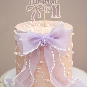 May include: A round, pale pink cake adorned with a large lavender bow and white pearl decorations. The cake has a topper that reads "Nimrah is 11". The cake is presented on a white and marble cake stand.