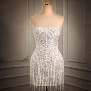 May include: A strapless white mini dress featuring an intricate pattern of silver sequins and beads, with a fringe hemline that hangs from the bottom. A gold-colored metal crown sits atop the mannequin head, completing the ensemble display.