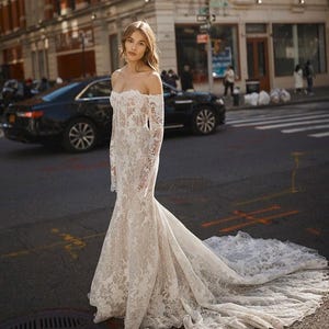 May include: A white, off-the-shoulder wedding dress with long sleeves and a flowing train. The dress is made of lace and is worn by a person standing in a city street with a brick building in the background.