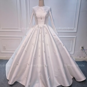 Puede incluir: Un vestido de novia de satén blanco con mangas largas y un escote redondo. El vestido tiene una falda completa y está decorado con delicados bordados florales.