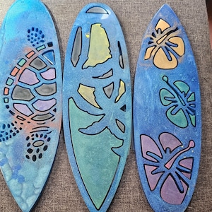 Puede incluir: Tres letreros de madera azules en forma de tabla de surf con diferentes diseños. El primer letrero tiene un diseño de tortuga colorido. El segundo letrero tiene un diseño de palmera colorido. El tercer letrero tiene un diseño de flor de hibisco colorido.