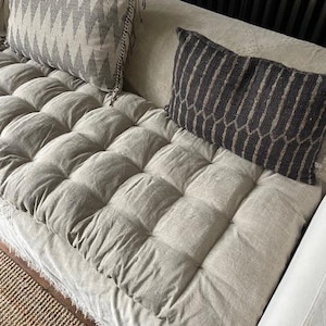 May include: A beige sofa with a tufted seat and decorative cushions. One cushion has a grey and white zigzag pattern, the other a dark grey textured design. The sofa's fabric looks soft and inviting, suitable for home decor. The sofa is in a home setting.