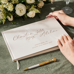 Pode incluir: Um livro de visitas de casamento creme com letras douradas que dizem "Christina and Adeline" e a data "Domingo, 10 de agosto de 2025". Uma caneta dourada com uma parte superior em forma de coroa repousa sobre uma toalha de mesa verde. Flores brancas no fundo.