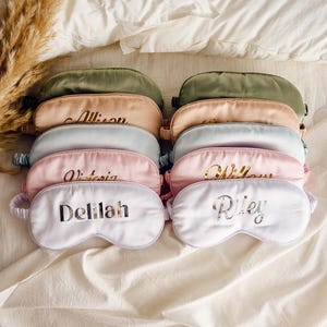 May include: A set of eight personalised sleep masks in various colours, including green, pink, and white. Each mask has a different name embroidered on it in gold lettering. The names are Allison, Victoria, Willow, Delilah, and Riley.