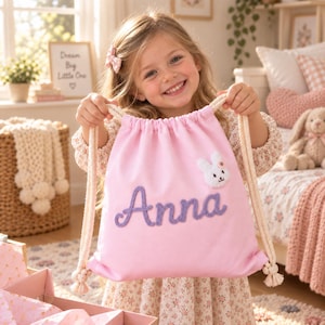 Puede incluir: Una mochila de cordón rosa claro con el nombre Anna en letras bordadas de color morado y una pequeña aplicación de peluche de conejo blanco en la parte superior derecha. La bolsa tiene correas de cuerda gruesa de color crema.