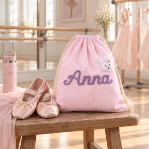 以下が含まれることがあります： 紫色の筆記体でAnnaと刺繍され、小さな白いウサギのワッペンが付いた薄いピンク色の巾着袋。袋の横には、淡いピンク色のサテン製バレエシューズ一足と、お揃いのリボン柄が描かれた薄いピンク色の水筒が置かれています。