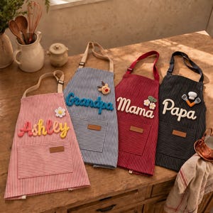 Könnte beinhalten: Vier gestreifte Schürzen mit Taschen und personalisierten Namen in erhabener Schrift. Die Schürzen sind rosa, blau, rot und schwarz und mit dekorativen Elementen wie einer Blume, einem Rentier und Küchenutensilien verziert.