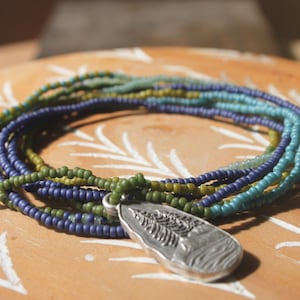May include: A multi-strand beaded necklace with a silver pendant. The necklace features strands of green, blue, and teal beads. The pendant is a silver teardrop shape with a detailed leaf design.