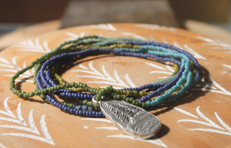 May include: A multi-strand beaded necklace with a silver pendant. The necklace features strands of green, blue, and teal beads. The pendant is a silver teardrop shape with a detailed leaf design.