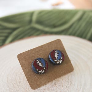 May include: A pair of wooden stud earrings with a red, white, and blue lightning bolt design. The earrings are on a brown card.