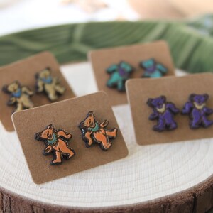 May include: Three pairs of earrings featuring a cartoon bear design. The earrings are displayed on brown card stock. The bears are wearing different colored clothing and have different colored fur. The earrings are a light blue, orange, and purple color.