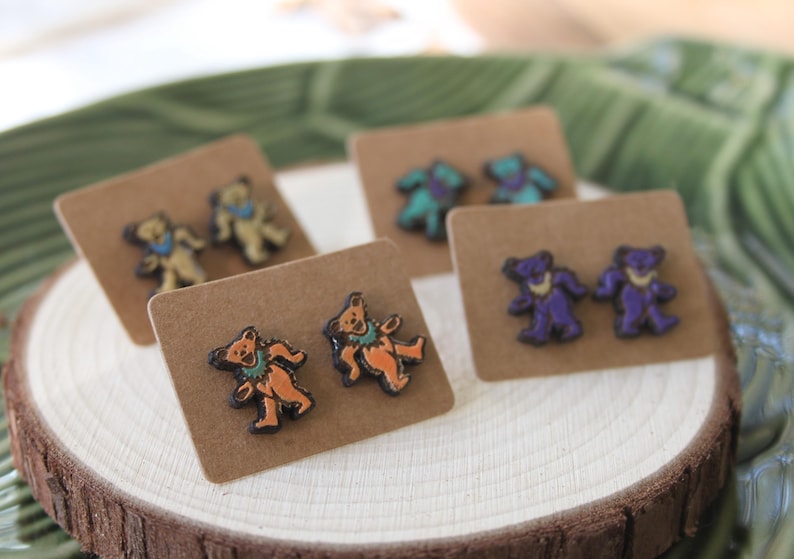 May include: Three pairs of earrings featuring a cartoon bear design. The earrings are displayed on brown card stock. The bears are wearing different colored clothing and have different colored fur. The earrings are a light blue, orange, and purple color.