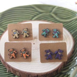 May include: Four pairs of wooden earrings featuring a dancing bear design. The bears are painted in different colors, including teal, orange, purple, and yellow.