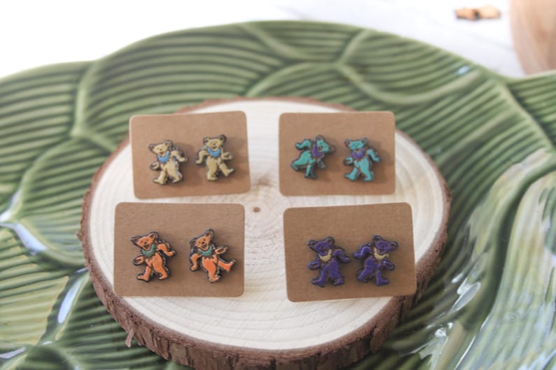 May include: Four pairs of wooden earrings featuring a dancing bear design. The bears are painted in different colors, including teal, orange, purple, and yellow.