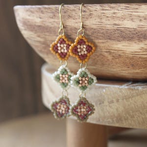 May include: Handmade beaded earrings with a geometric design. The earrings feature a gold-colored hook and three diamond-shaped sections. The top section is orange and maroon, the middle section is green and white, and the bottom section is maroon and copper.