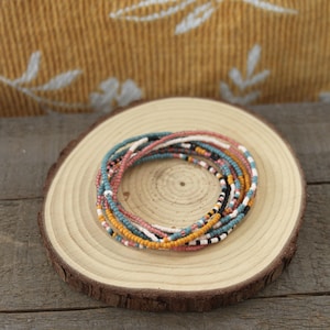 May include: A collection of colorful beaded bracelets in shades of pink, blue, yellow, black, and white. The bracelets are arranged in a circular pattern on a wooden slice, set against a rustic wooden background.