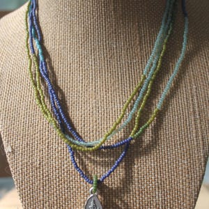 May include: A multi-strand beaded necklace in shades of blue, green, and teal. The necklace features a silver pendant with a detailed tree and landscape design. The necklace is displayed on a burlap-covered bust.