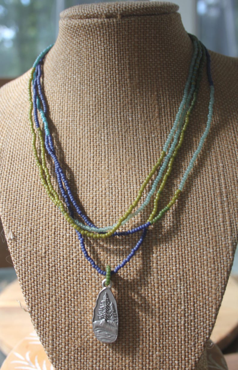 May include: A multi-strand beaded necklace in shades of blue, green, and teal. The necklace features a silver pendant with a detailed tree and landscape design. The necklace is displayed on a burlap-covered bust.