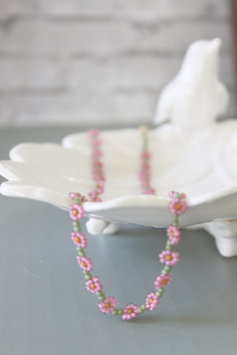 May include: A pink and green beaded necklace with small flower-shaped beads. The necklace is displayed on a white ceramic dish.
