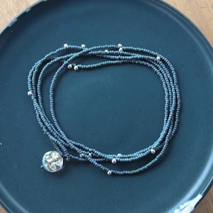 以下が含まれることがあります： 小さな濃い青色のビーズと銀色のアクセントでできたマルチストランドネックレス。ネックレスには丸い虹色のペンダントが付いています。