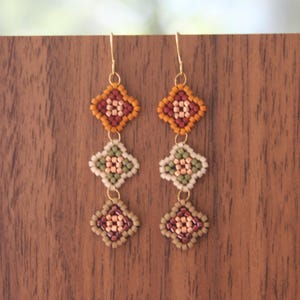 May include: Handmade beaded earrings with a geometric design. Each earring features three diamond-shaped sections in shades of orange, red, green, and beige, suspended from gold-colored hooks. The earrings are displayed against a wooden background.