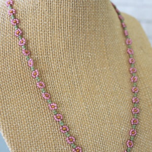 May include: A delicate pink and green beaded necklace with small flower-shaped beads. The necklace is displayed on a brown fabric mannequin.