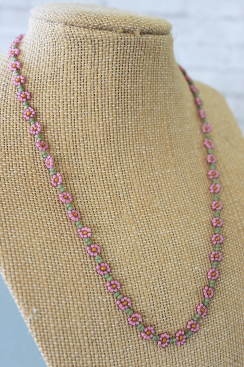 May include: A delicate pink and green beaded necklace with small flower-shaped beads. The necklace is displayed on a brown fabric mannequin.