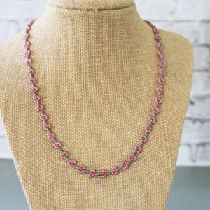 May include: A delicate pink and green beaded necklace with small flower-shaped beads. The necklace is displayed on a brown fabric mannequin.