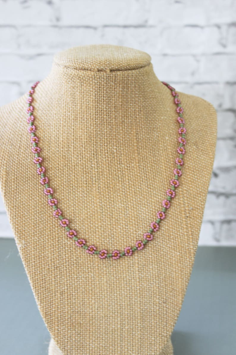 May include: A delicate pink and green beaded necklace with small flower-shaped beads. The necklace is displayed on a brown fabric mannequin.