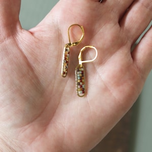 Puede incluir: Un par de pendientes de aro dorados con un diseño de cuentas de colores. Las cuentas son en tonos de marrón, rojo, blanco y beige.