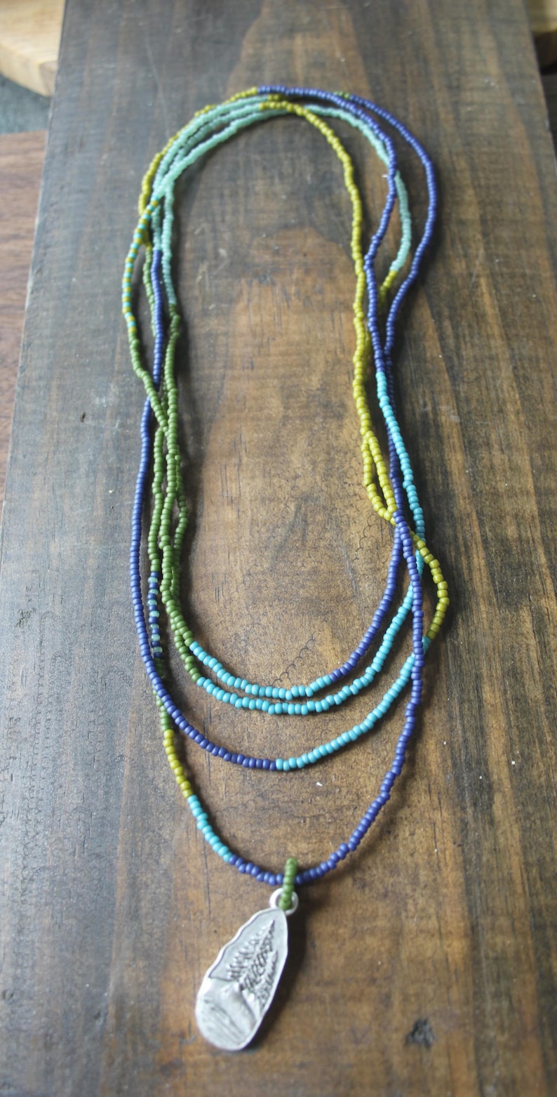 May include: A multi-strand necklace featuring small beads in blue, green, and yellow. A silver pendant with a tree design is attached to the lowest strand. The necklace is presented on a wooden surface.