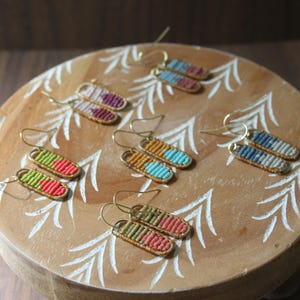 May include: A collection of handmade macrame earrings in various colors, including red, green, blue, and yellow. Each pair features a woven design with gold-colored ear wires. The earrings are displayed on a decorative wooden stand.