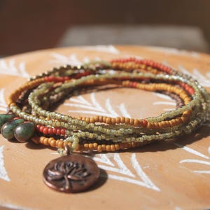 May include: A multi-strand beaded necklace in shades of green, orange, and brown, with a copper-colored pendant featuring a tree design. The necklace is resting on a wooden surface with a white leaf pattern.