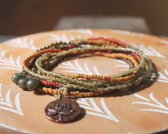 May include: A multi-strand beaded necklace in shades of green, orange, and brown, with a copper-colored pendant featuring a tree design. The necklace is resting on a wooden surface with a white leaf pattern.
