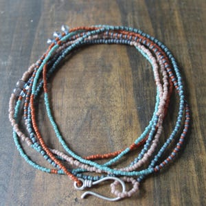 May include: A multi-strand beaded necklace in shades of teal, rust, and tan. The necklace features small, round beads strung together, with a silver-colored hook clasp. The jewelry is displayed on a wooden surface.