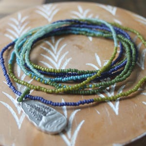 May include: A beaded necklace with a silver pendant rests on a wooden surface. The necklace features strands of small, colourful beads in shades of green, blue, and purple. The pendant has a detailed design.