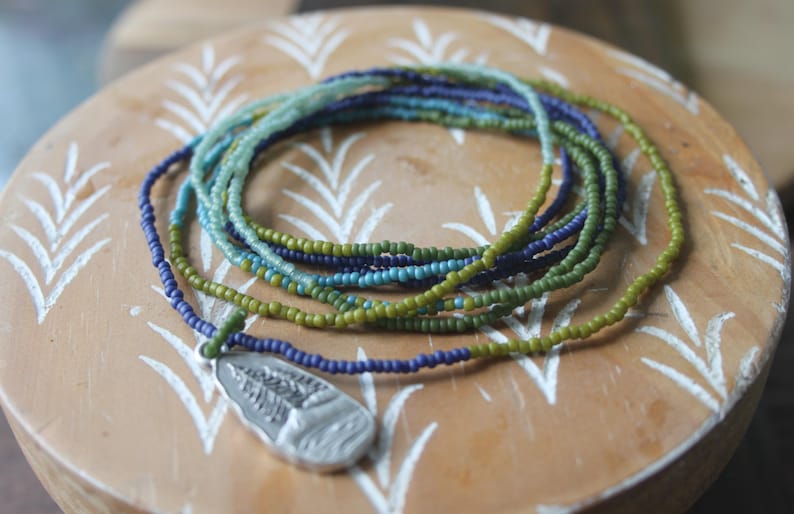 May include: A beaded necklace with a silver pendant rests on a wooden surface. The necklace features strands of small, colourful beads in shades of green, blue, and purple. The pendant has a detailed design.