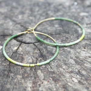May include: A pair of gold hoop earrings with green and yellow beads. The beads are arranged in a gradient pattern, starting with a light green at the top and fading to a darker green at the bottom.