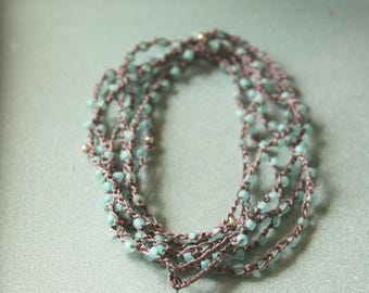 May include: A handmade necklace with multiple strands of brown thread and small, light blue beads. The necklace has a flower pendant with a light blue and pink design. The jewelry is displayed on a light blue surface.
