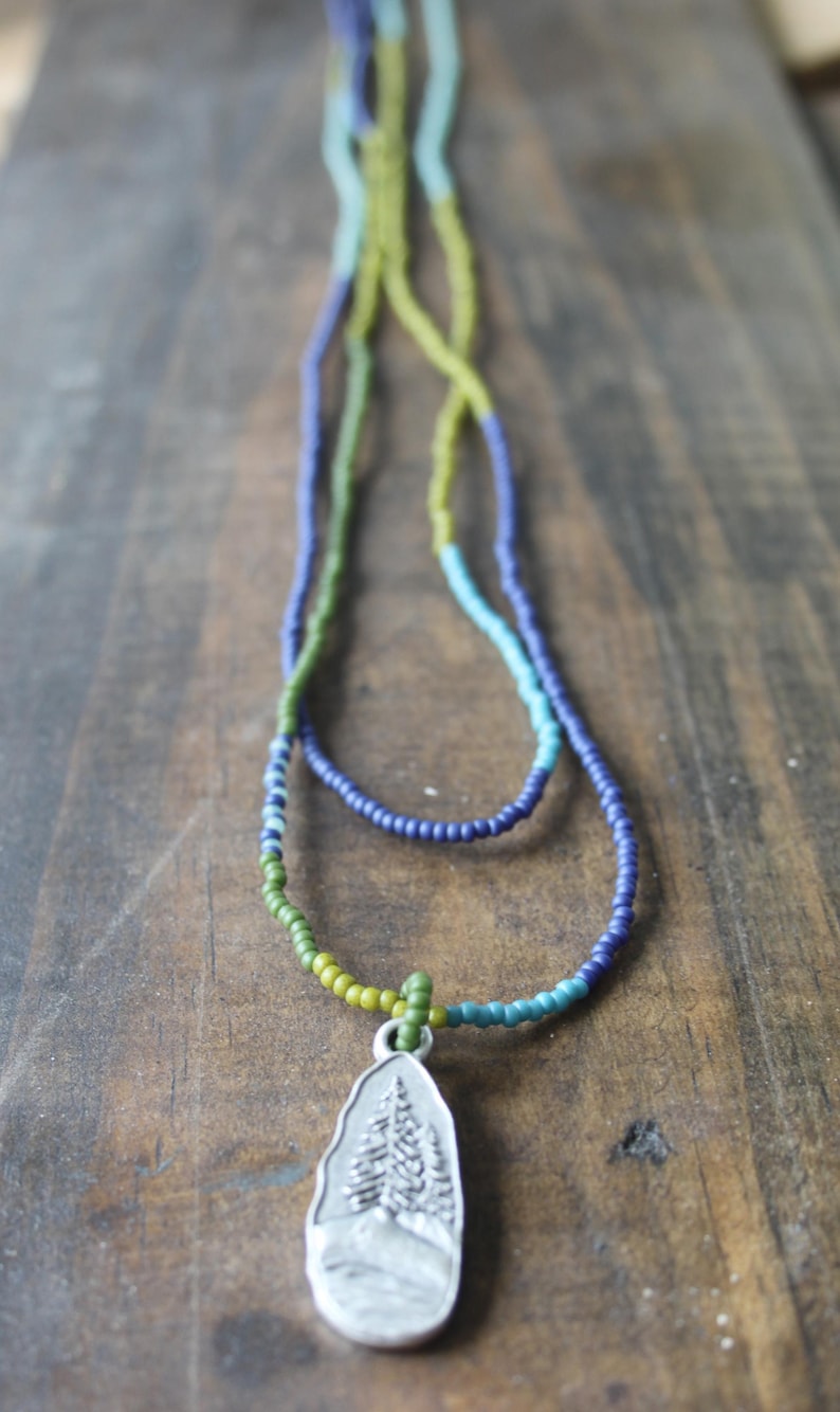 May include: A necklace with a silver pendant and strands of green, blue, and yellow beads. The pendant is a silver teardrop shape with a mountain and tree design. The necklace is displayed on a wooden surface.
