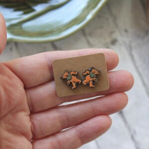 May include: A pair of brown wooden earrings with a design of two dancing bears. The earrings are on a small brown card.