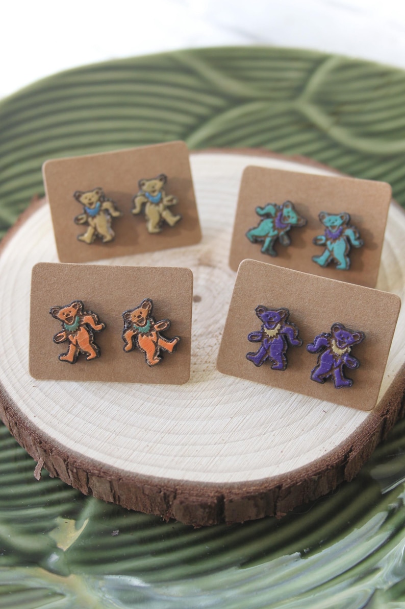May include: Four pairs of wooden earrings featuring a dancing bear design. The bears are painted in different colors, including orange, purple, blue, and yellow.