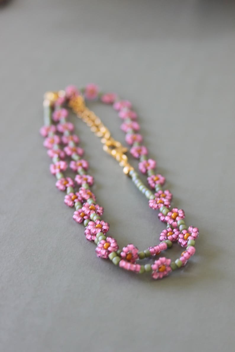 May include: A delicate double-strand necklace with pink flower-shaped beads and green accents. The necklace is made of small beads and has a gold clasp.
