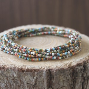 Op de afbeelding: Een verzameling kleurrijke kralenarmbanden, met kleine, ronde kralen in verschillende kleuren, waaronder blauw, groen, oranje en wit. De armbanden zijn in een cirkelvorm op een rustiek houten oppervlak gerangschikt.