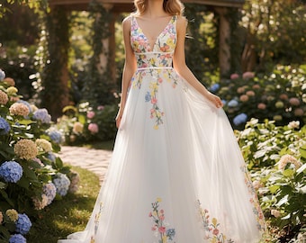Puede incluir: Vestido de novia blanco sin mangas con escote en V y bordado floral. El vestido presenta una falda fluida y un delicado estampado floral en tonos rosa, amarillo y azul. El vestido se usa al aire libre en un jardín.
